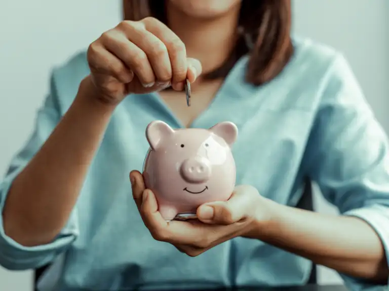 Mãos de uma mulher vestindo camisa azul claro depositando uma moeda em um cofrinho de porco rosa
